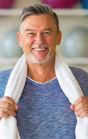 A man is seen in a gym setting, holding a towel around his neck.