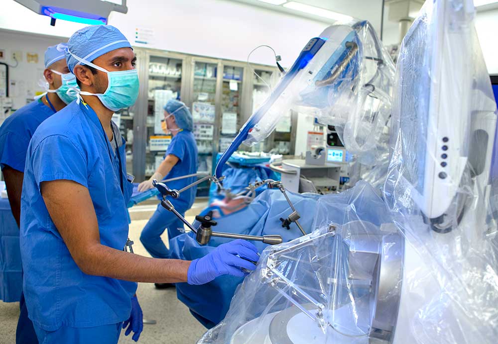 Dr. Neerav Goyal looks at a monitor while using the Medrobotics Flex System to perform head and neck surgery on a patient at Milton S. Hershey Medical Center. Behind him are a man and woman. All three are wearing hospital scrubs, surgical masks and gloves. The patient is lying on a gurney with two silver instruments attached to his or her mouth and is covered with blue plastic. The surgical equipment is covered in clear plastic.