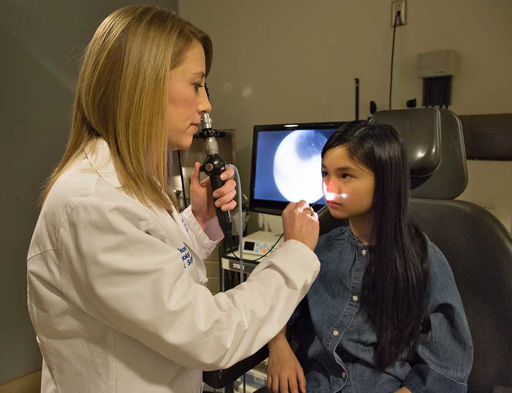 Dr. Meghan Wilson examines a young girl using a rhinolaryngoscope. Dr. Wilson wears a white physician’s coat and holds the handle of the tool in one hand and aims the small camera tube up the patient’s nose with the other. The patient has long, dark hair and is looking at Wilson while sitting upright in an examination chair. The video picture created by the instrument’s light can be seen on a monitor in the background.