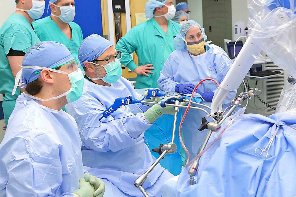 Surgeon uses Flex robot in operating room surrounded by staff. Staff and surgeon in green and light blue scrubs. Equipment directly in front of surgeon.