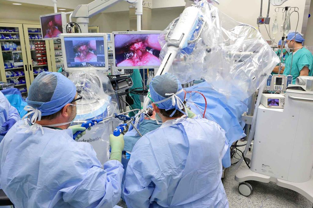 Operating room with surgeon using Flex robot to remove head and neck cancer. Staff are in light blue scrubs. Three screens show the operation.