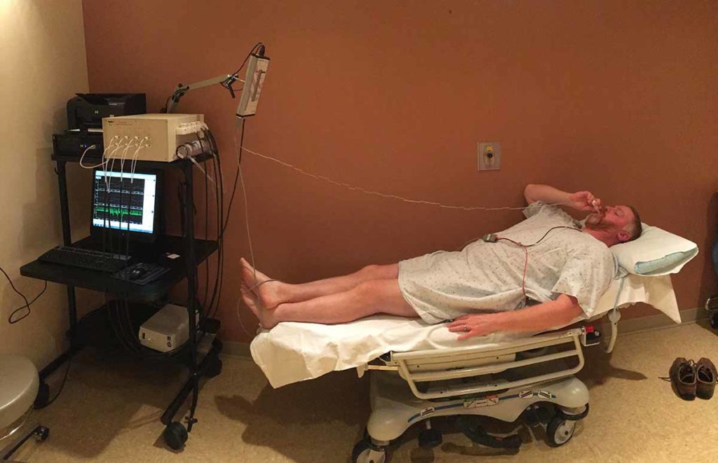 A patient in a gown on a hospital bed is next to equipment with lights and wires. The equipment is for the Quantitative Sudomotor Axon Reflex Test or QSART and Vasalva maneuver tests in the autonomic testing lab.