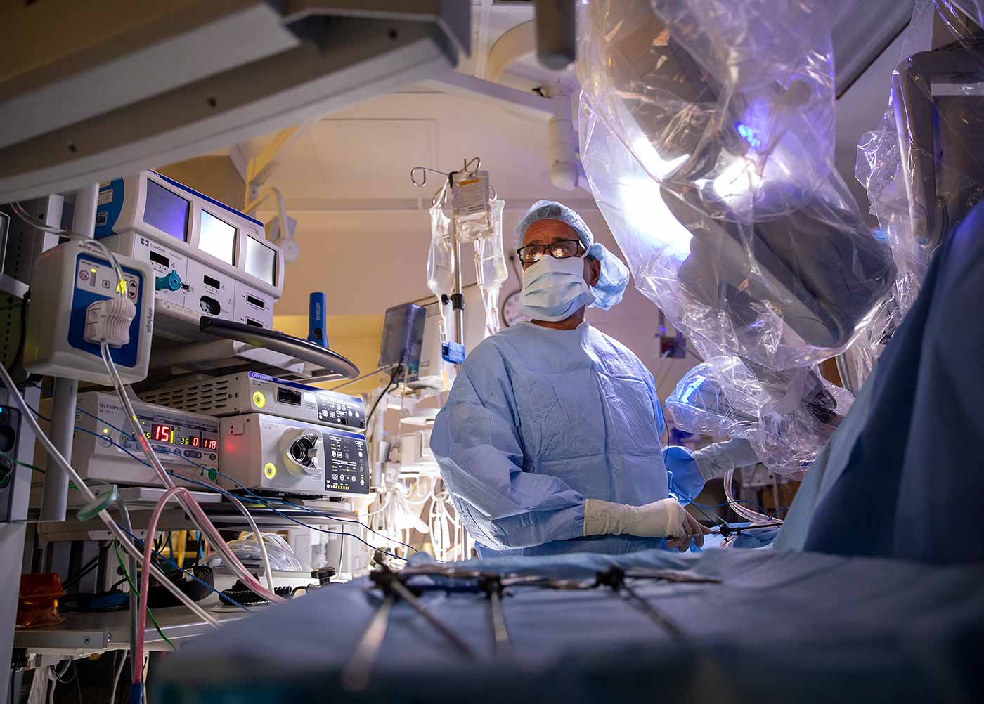 Dr. Jay Raman is in the operating room. He is in a blue gown, mask and cap. There is medical equipment in the background and medical instruments in the foreground.